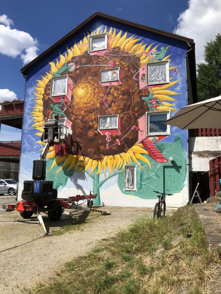 En bild från ett event från en Nässelfrossavecka. Konstnären och målaren Kim Demåne, målar en hel husgavel vid tre tillfällen i realtid under veckan. Huset lyser nu upp gatan för alla som färdas på denna in- och utfartsväg inne i Olofström.